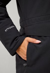 Zwarte waterdichte jas met een ritszak en geborduurd logo op de mouw. De stof heeft een gladde, matte textuur.