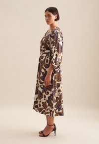 Mittelkleid mit floralen Mustern in Cremetönen, Marineblau und Gold, mit V-Ausschnitt und 3/4-Ärmeln. Kombiniert mit schwarzen High Heels.