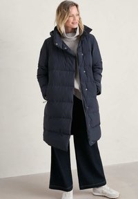 Larga chaqueta de plumas azul marino con capucha, que presenta segmentos acolchados y una cremallera frontal. Combinada con un suéter ligero y pantalones de pierna ancha oscuros.