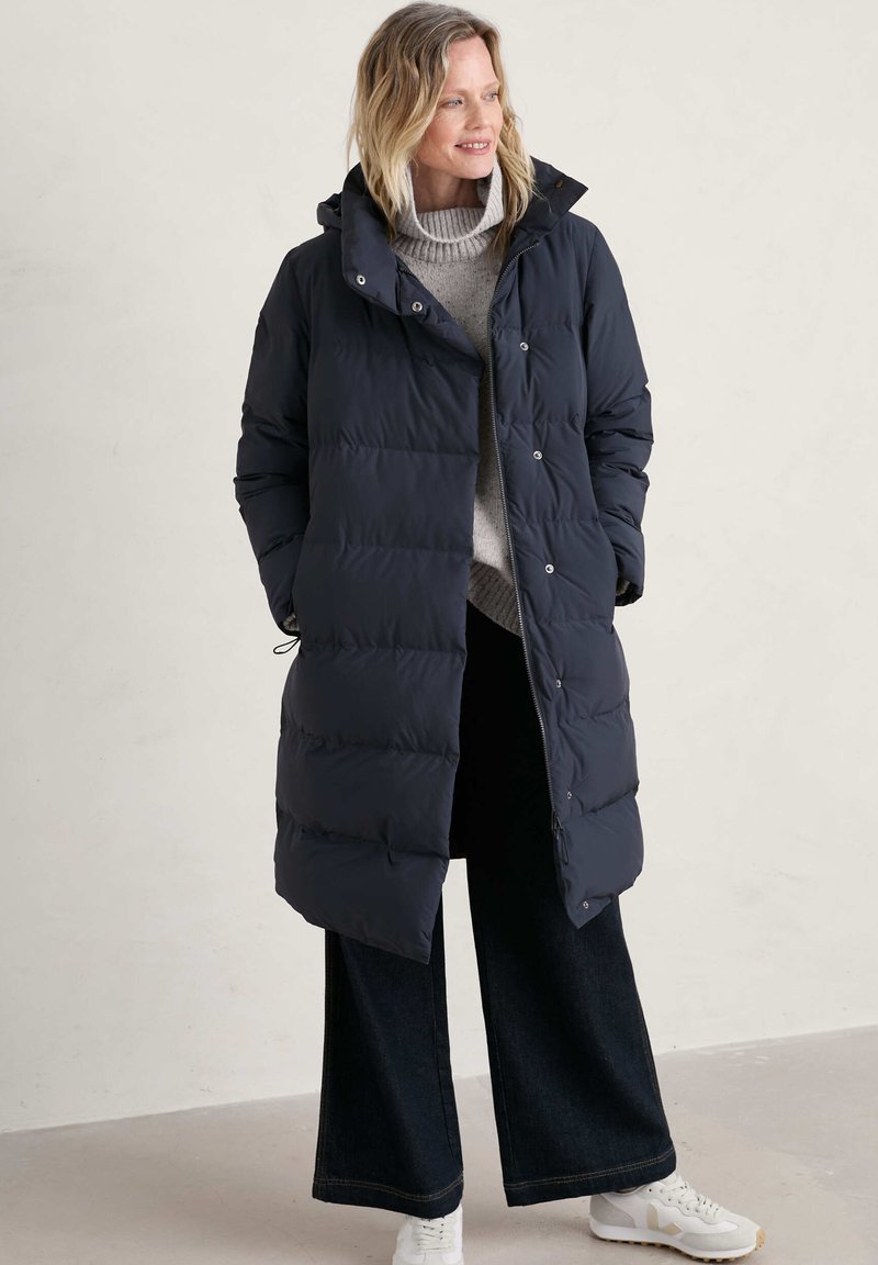 Larga chaqueta de plumas azul marino con capucha, que presenta segmentos acolchados y una cremallera frontal. Combinada con un suéter ligero y pantalones de pierna ancha oscuros.