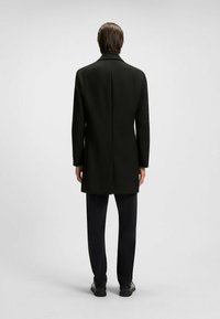 Uomo con capelli castani lisci che indossa un lungo cappotto nero, pantaloni neri e scarpe nere, in piedi con le spalle rivolte verso di noi su uno sfondo chiaro uniforme.