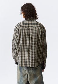 Chemise à carreaux beige et marine avec une coupe décontractée, dotée de manches longues, d'un pli au dos et d'un ourlet arrondi. Un pantalon en denim est porté.