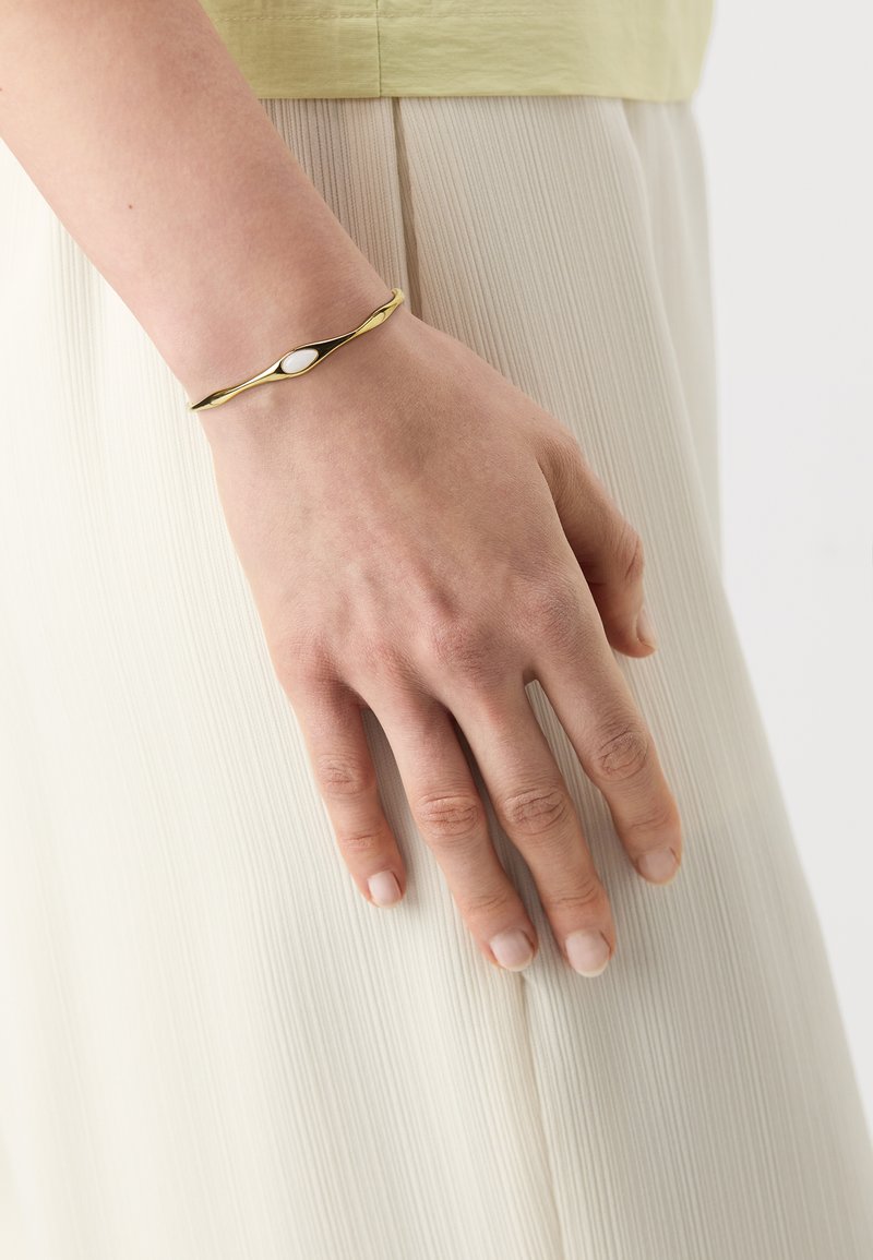 Pulsera de oro con un diseño retorcido que presenta un acento ovalado blanco, llevada en una mano frente a un fondo de tela clara y plisada.