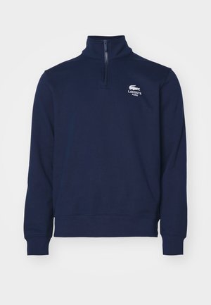 Pulover bleumarin din bumbac cu guler înalt, fermoar până la jumătate, mansete cu nervuri și un logo brodat alb pe partea stângă a pieptului. Textură de țesătură moale.