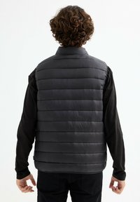 Chaleco de plumón negro con acolchado horizontal, cuello alto y una textura lisa y brillante, usado sobre una camiseta negra de manga larga.