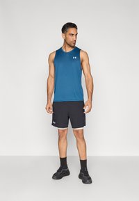 Top sportivo senza maniche blu abbinato a pantaloncini neri con logo. La modella indossa scarpe da ginnastica nere e calzini neri, in piedi contro uno sfondo semplice.