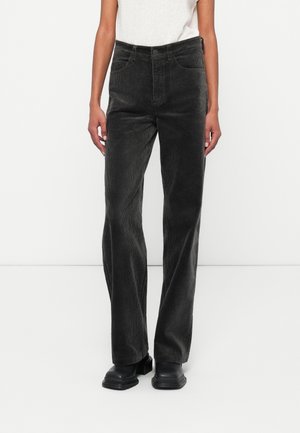 Vrouw draagt zwarte wijde ribfluwelen broek met hoge taille, wit gebreid mouwloos topje en zwarte stevige laarzen, staand tegen een witte achtergrond.