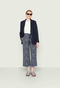 Blazer de linho azul-marinho com dois botões, t-shirt branca, calças largas estampadas e sandálias cinzas. Óculos de sol e brincos de argola incluídos.
