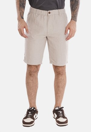 Bermuda in lino beige con una vestibilità rilassata, cintura elasticizzata e dettaglio dei bottoni. Indossate con sneaker marroni e bianche, che mostrano un tessuto testurizzato.