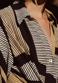 Kragenhemd mit schwarzen, beigen und weißen vertikalen sowie horizontalen Streifen, getragen von einer Person mit kurzen blonden Haaren, teilweise sichtbarem Hals und Schulter.