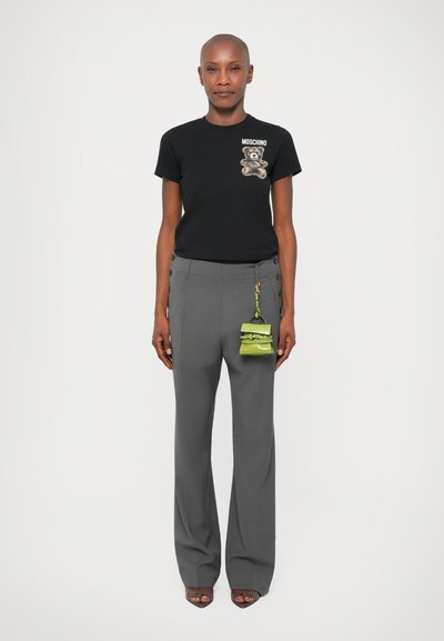 Mulher a usar uma t-shirt preta da Moschino com estampa de ursinho, calças largas de cintura alta cinzentas com botões e uma pequena mala verde presa à cintura.