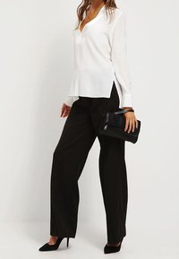 Blusa bianca a maniche lunghe con scollatura a V e spacchi laterali, abbinata a pantaloni neri a gamba larga a vita alta. Borsa clutch nera e tacchi.