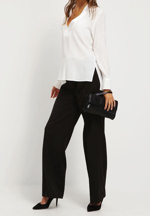 Blusa bianca a maniche lunghe con scollatura a V e spacchi laterali, abbinata a pantaloni neri a gamba larga a vita alta. Borsa clutch nera e tacchi.