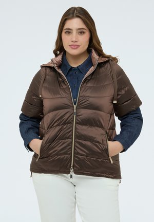 Młoda kobieta w brązowej krótkiej pikowanej kurtce na niebieskiej dżinsowej koszuli i białych spodniach, stojąca z rękami w kieszeniach kurtki.