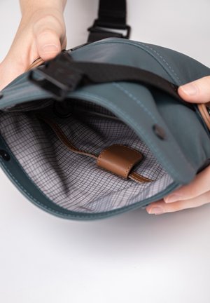 Innenraum einer türkisen Ledertasche mit einem Innenfutter im Karomuster. Außen mit einem braunen Lederriemen und einem verstellbaren schwarzen Riemen ausgestattet.