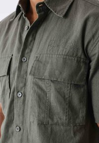 Camisa de manga corta verde hecha de tela texturizada, con dos bolsillos en el pecho, cierre de botones y un logo bordado en el bolsillo izquierdo.