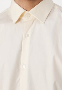 Camicia a maniche corte color crema con bottoni. Presenta un colletto classico, una texture del tessuto liscia e due bottoni visibili nella zona del collo.