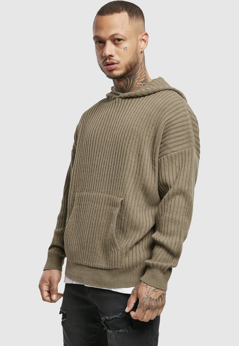 Homme avec des tatouages au cou et aux mains, portant un sweat à capuche en maille côtelée olive et un jean noir déchiré, debout devant un fond gris clair.