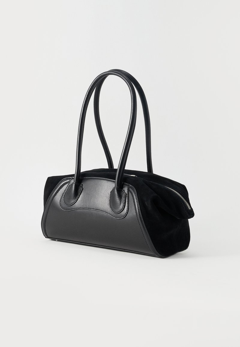 Black handbag with long rounded handles, leather front panel, and a zippered top, standing upright on a plain white background.