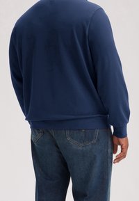 Uomo che indossa una felpa a maniche lunghe blu navy e jeans blu scuro, in piedi con la schiena rivolta verso uno sfondo neutro.