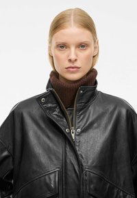 Veste en cuir noir avec un col montant, une fermeture éclair et des pressions. Un col roulé côtelé marron est porté en dessous.