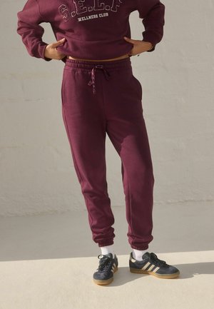 Burgundy sweatpants with an elastic waistband and drawstring, paired with black sneakers featuring beige accents and white socks.