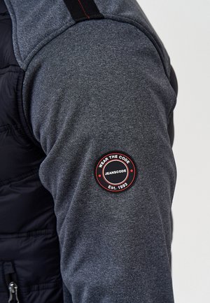 Grå sweatshirtstoff med svart vadderad sektion, med en rund svart och röd logotyp på ärmen. Slät textur, avslappnad design.