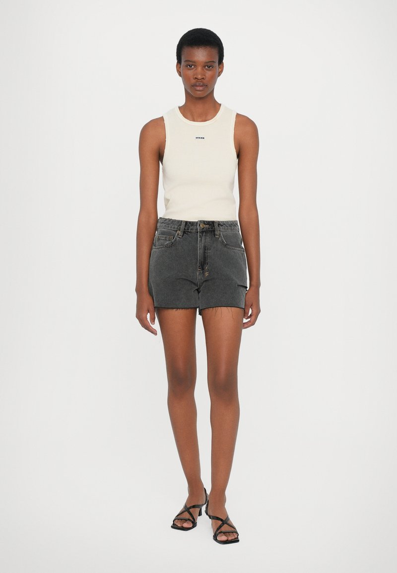 Canottiera a coste beige chiaro, pantaloni corti in denim neri a vita alta e sandali neri con fasce. Il modello è in piedi con le braccia rilassate ai lati.