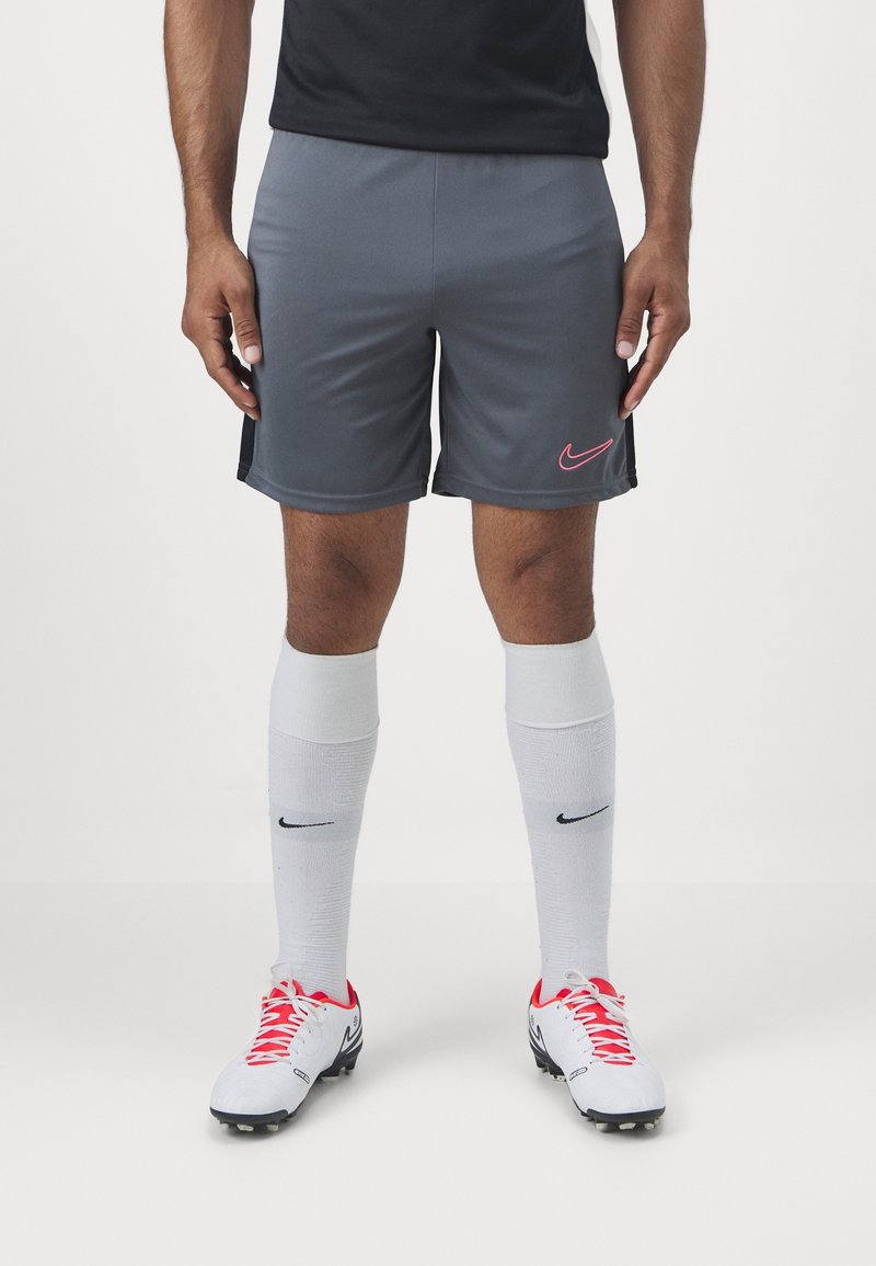 Shorts de sport gris avec un logo Nike rose, chaussettes blanches montantes avec un logo noir, et chaussures blanches avec lacets rouges et accents noirs.