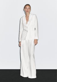 Donna in piedi su uno sfondo bianco, vestita con un completo bianco largo composto da pantaloni a gamba larga e giacca, che guarda dritto di fronte a sé con un'espressione neutra.