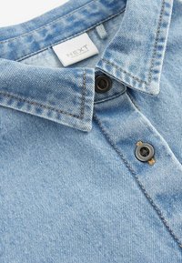 Chemise en denim bleu clair avec un col pointu, des boutons noirs et des coutures contrastées. L'étiquette indique "NEXT, FABRIQUÉ EN INDE." Texture lisse.