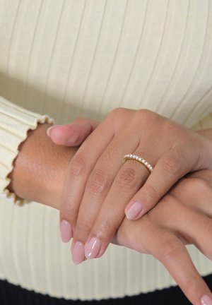 Goldring mit kleinen weißen Edelsteinen, getragen an einer Hand mit zart rosa Nagellack. Die Hände sind zusammengelegt, im Hintergrund ist ein strukturiertes cremefarbenes Oberteil zu sehen.