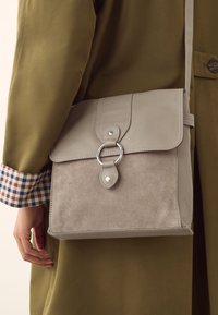 Hall hall käekott, mille esiküljel on tekstuuriga seemisnahk, tagaküljel sile nahk, ümmargused metallist detailid ja klapiga kinnitamine. Kaasas reguleeritav rihm.