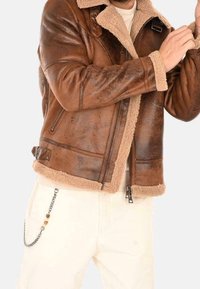 Mężczyzna w brązowej kurtce ze skórzanym barankiem z suwakiem i detalem z klamrą, ubrany w jasno-beżowe spodnie z łańcuszkową ozdobą.