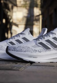Chaussures de running blanches Adidas avec maille noire mouchetée, trois bandes noires, lacets à motifs et texte « Light Boost » sur la semelle amortie.