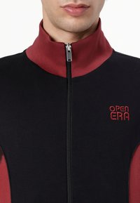 Giacca con zip nera e bordeaux con collo alto, caratterizzata da ricamo del logo e tessuto texture, con un design elegante e aderente.