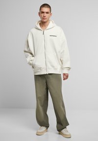 Hvid zip-up hoodie med sidelommer, manchetter og sort logo. Kombineret med olivengrønne bukser og cremede sneakers. Glat stofstruktur.