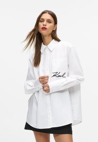 Kvinne med langt brunt hår iført en oversized hvit skjorte med "Karl" broderi på lommen og svart skjørt mot hvit bakgrunn.