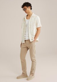Camisa de manga corta color crema con patrón floral marrón, camiseta sin mangas blanca, pantalones ajustados color beige y zapatos slip-on color beige. Unas gafas de sol a la moda completan el look.