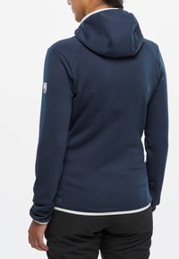 Granatowa polarowa kurtka z kapturem o gładkiej fakturze, dopasowanym kroju i białym wykończeniu na dole. Logo na lewym rękawie.