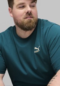 Mėlynas trumpas rankovėmis marškinėliai, pagaminti iš minkštos medžiagos, su kreminiu siuvinėtu Puma logotipu ant krūtinės ir apvaliu iškirpimu.