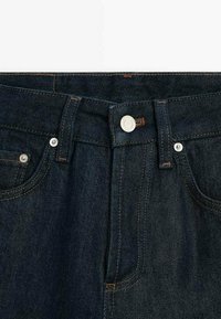Jean en denim bleu foncé à la taille avec bouton argenté, passants de ceinture, poches avant et coutures orange sur fond blanc.