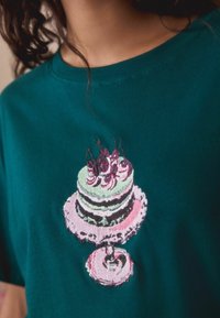 Turkusowa bawełniana koszulka z haftowanym wzorem tortu, który ma zielone i różowe warstwy, udekorowany wiśniami i bitą śmietaną, na stojaku na ciasto.