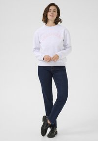Lichtgrijze sweatshirt met "GRATITUDE" in roze letters over de borst, gecombineerd met donkerblauwe jeans en zwarte schoenen. Glad textuur.