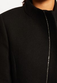 Zwarte jas met een textuurstof, hoge kraag en een decoratieve zilveren ritsaccent langs de voorkant.