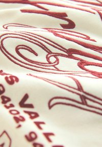 Wyszywana tkanina z kremowym tłem i bogatym burgundowym tekstem, z falami i wyraźnym pismem, prezentująca gładką teksturę.