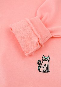 Felpa rosa con polsini testurizzati e un design di gatto ricamato in nero e verde sulla parte anteriore. Tessuto in cotone morbido.