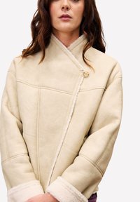 Vrouw in een crèmekleurige schapenvachtjas met een knoopsluiting en een opstaande kraag, zichtbaar van borst tot kin.