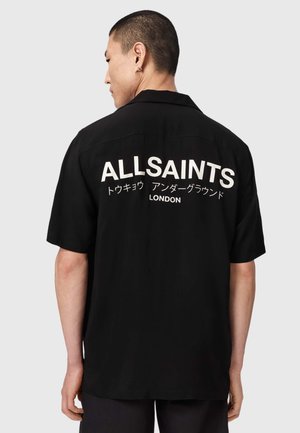 Man met kort haar die een zwart shirt met korte mouwen draagt met witte tekst "ALLSAINTS Tokyo Underground London" op de rug.