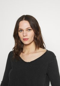 Chemise noire à manches longues avec un col en V, présentant un col légèrement effiloché et une texture douce. Le tissu semble décontracté et confortable.